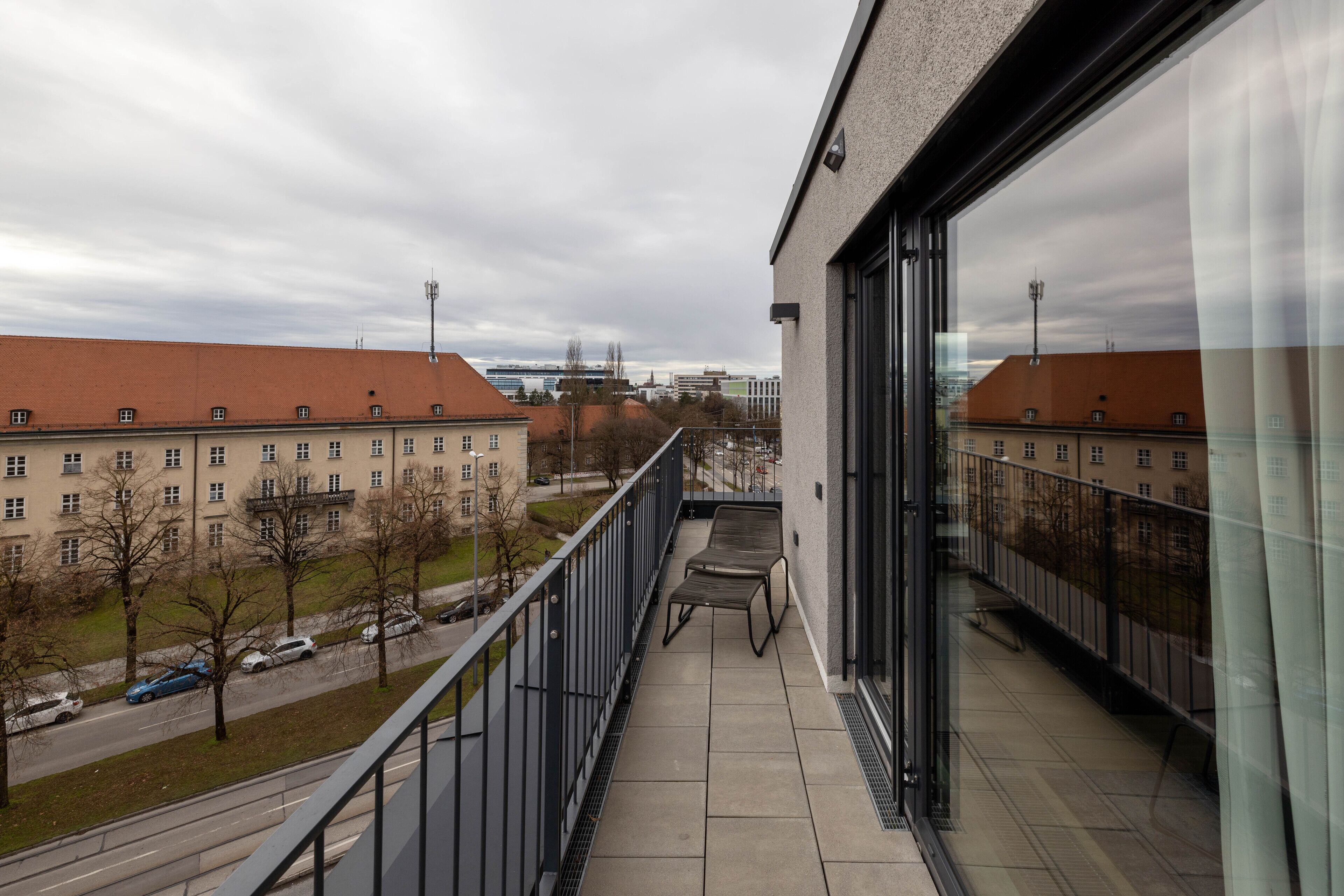 A balcony with a view of a building with a red roof, a paved walkway, and a metal railing.
