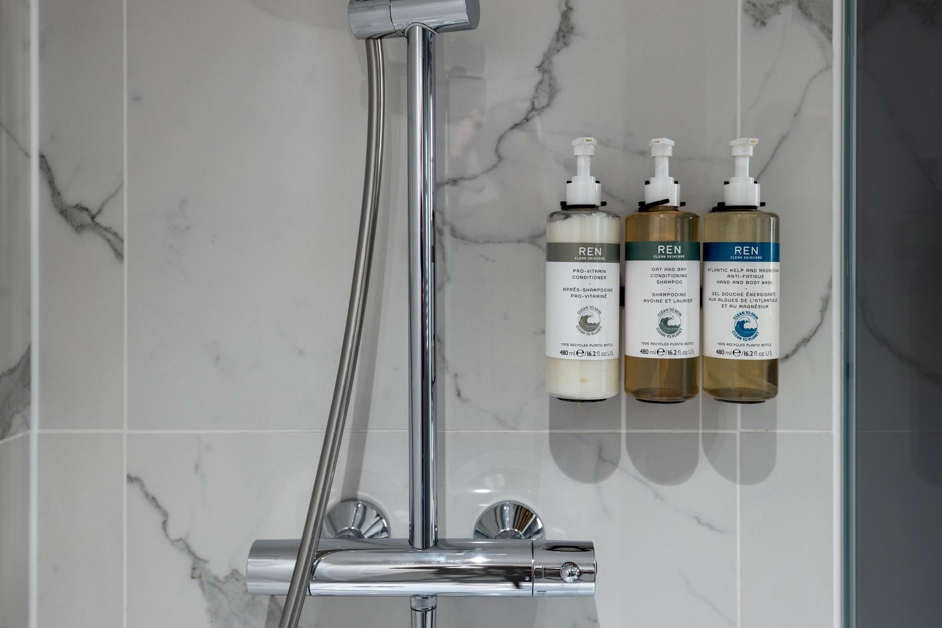 A bathroom with a shower, a hand dryer, and three bottles of REN body wash.