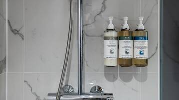A bathroom with a shower, a hand dryer, and three bottles of REN body wash.