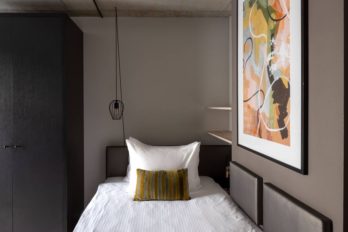 Een net opgemaakt bed met witte lakens en een gestreept kussen, een donkere kast en een ingelijst abstract schilderij aan de muur.