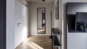 A hallway with a wooden floor, a white door, a mirror, a black floor lamp, and a black TV stand.