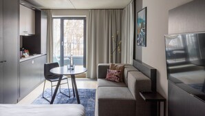 A modern hotel room with a sofa, a dining table, a TV, and a kitchenette.