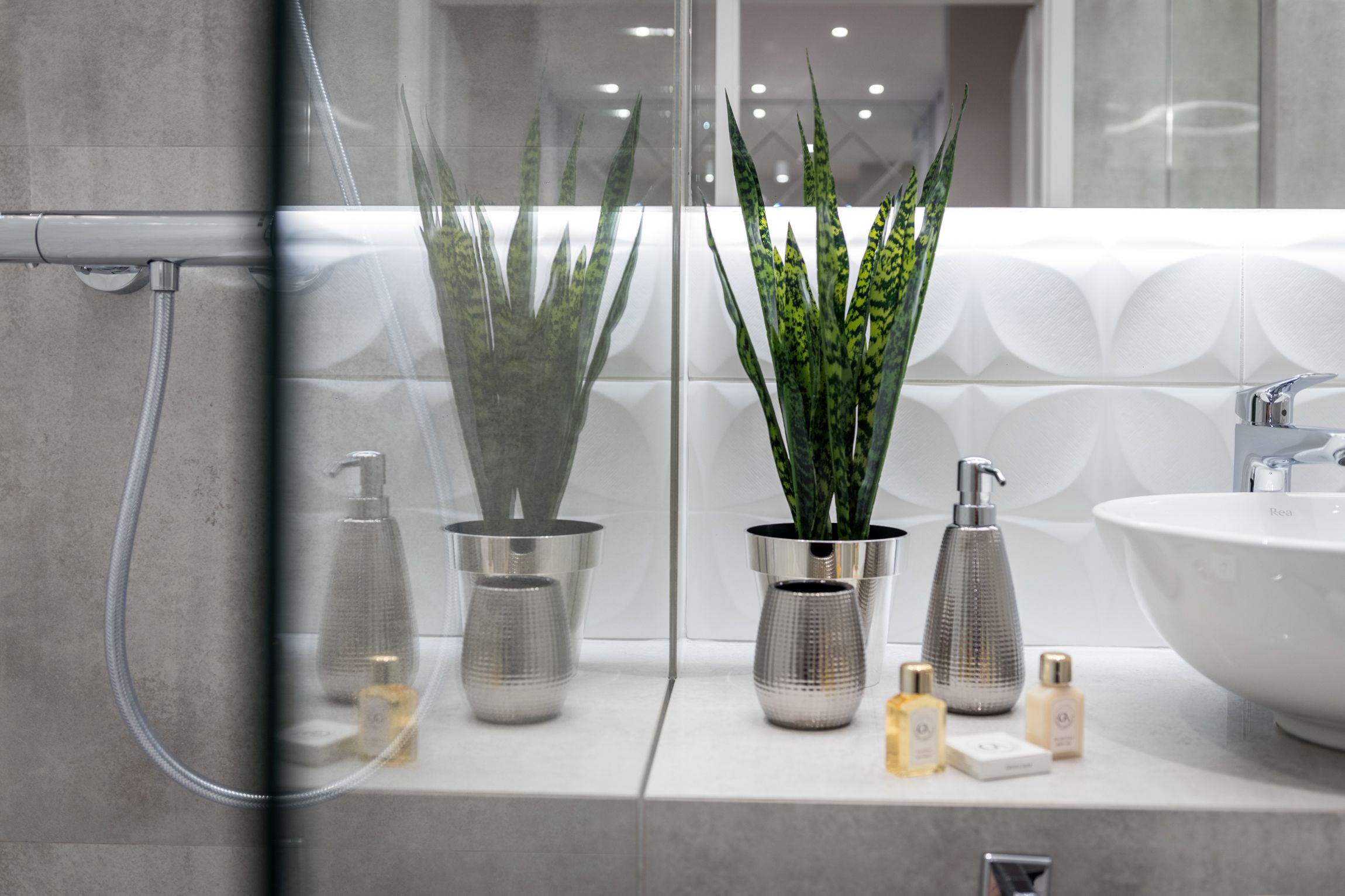 Un baño moderno con un espejo grande, dos plantas en macetas, una ducha y un lavabo con artículos de aseo.