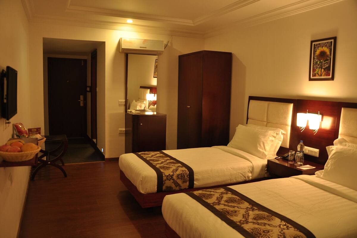 Quarto de hotel com duas camas, um guarda-roupa, uma secretária e uma pequena mesa com abajur.
