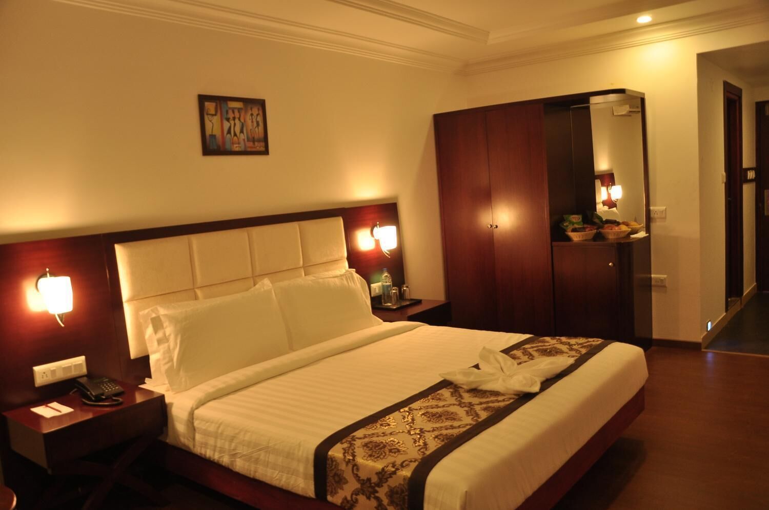 A hotel room with a large bed, a wooden wardrobe, a bedside table with a lamp, and a wall-mounted artwork.