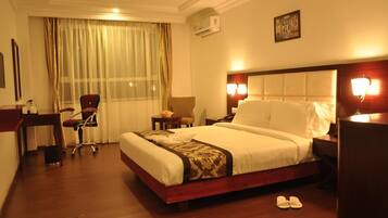 A hotel room with a large bed, a desk, a chair, a nightstand, and a wall-mounted air conditioner.
