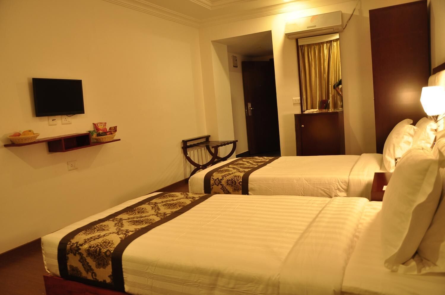 A hotel room with two beds, a TV mounted on the wall, a small table with a basket of fruit, and a chair.
