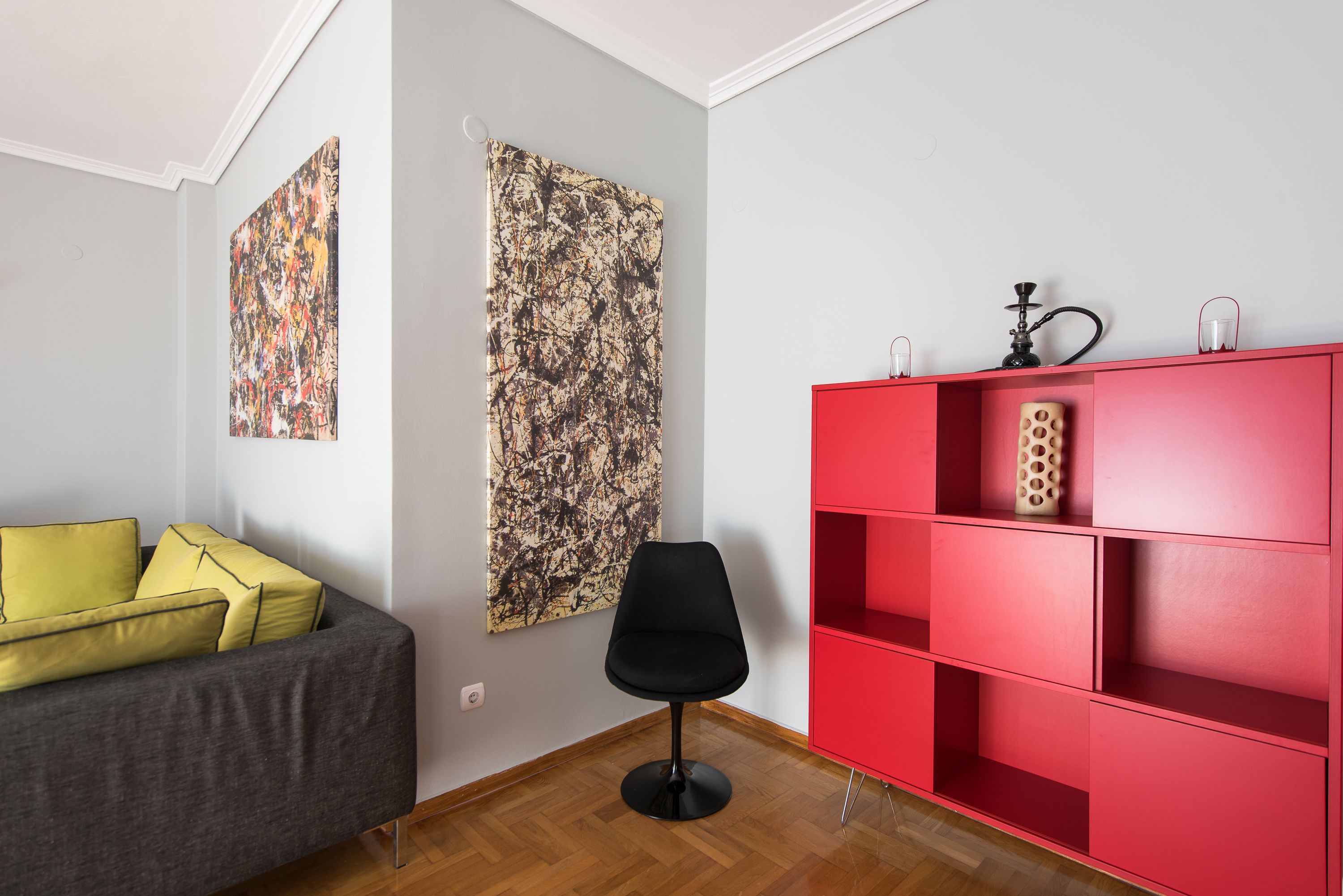 A modern living room with a grey sofa, yellow cushions, a black chair, and a red shelving unit against a white wall.