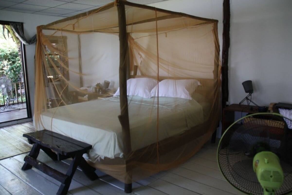 A four-poster bed with a mosquito net, a wooden bench, and a green fan.