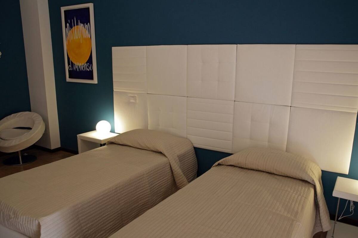 A hotel room with two beds, a white headboard, a bedside table with a lamp, and a wall-mounted artwork.