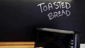 Un grille-pain posé sur une table en bois, avec deux tranches de pain sur une assiette, et les mots « Pain grillé » écrits sur un tableau noir en arrière-plan.