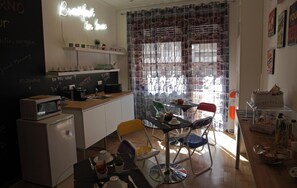 Una cucina compatta con forno a microonde, tostapane e macchina del caffè. Presente una lavagna con proposte di menu e un neon. La stanza ha una tenda con motivo cittadino e un tavolo con sedie.