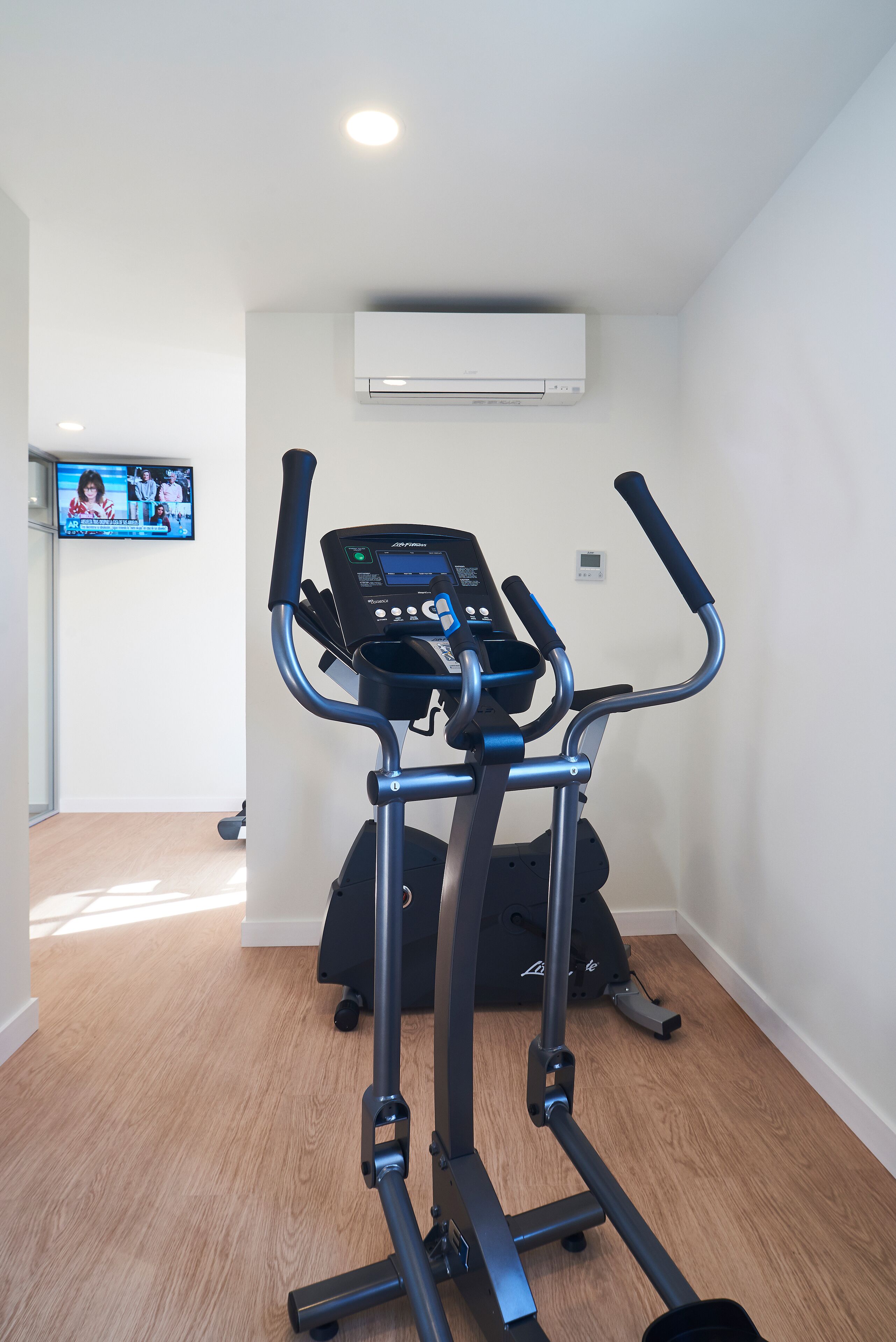 Une salle de sport à domicile équipée d’un tapis de course et d’un vélo elliptique, d’un téléviseur fixé au mur et d’un climatiseur installé au plafond.