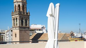 A rooftop terrace with two sun loungers and a white umbrella, overlooking a historic tower and cityscape. - Slow Suites Setas (Seville)