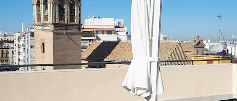 A rooftop terrace with two sun loungers and a white umbrella, overlooking a historic tower and cityscape.