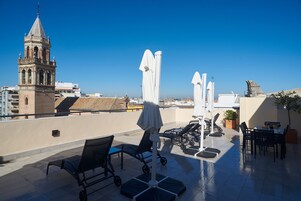 Terraza en la azotea con vistas a una torre, sillones de descanso, sombrillas y un área de comedor.