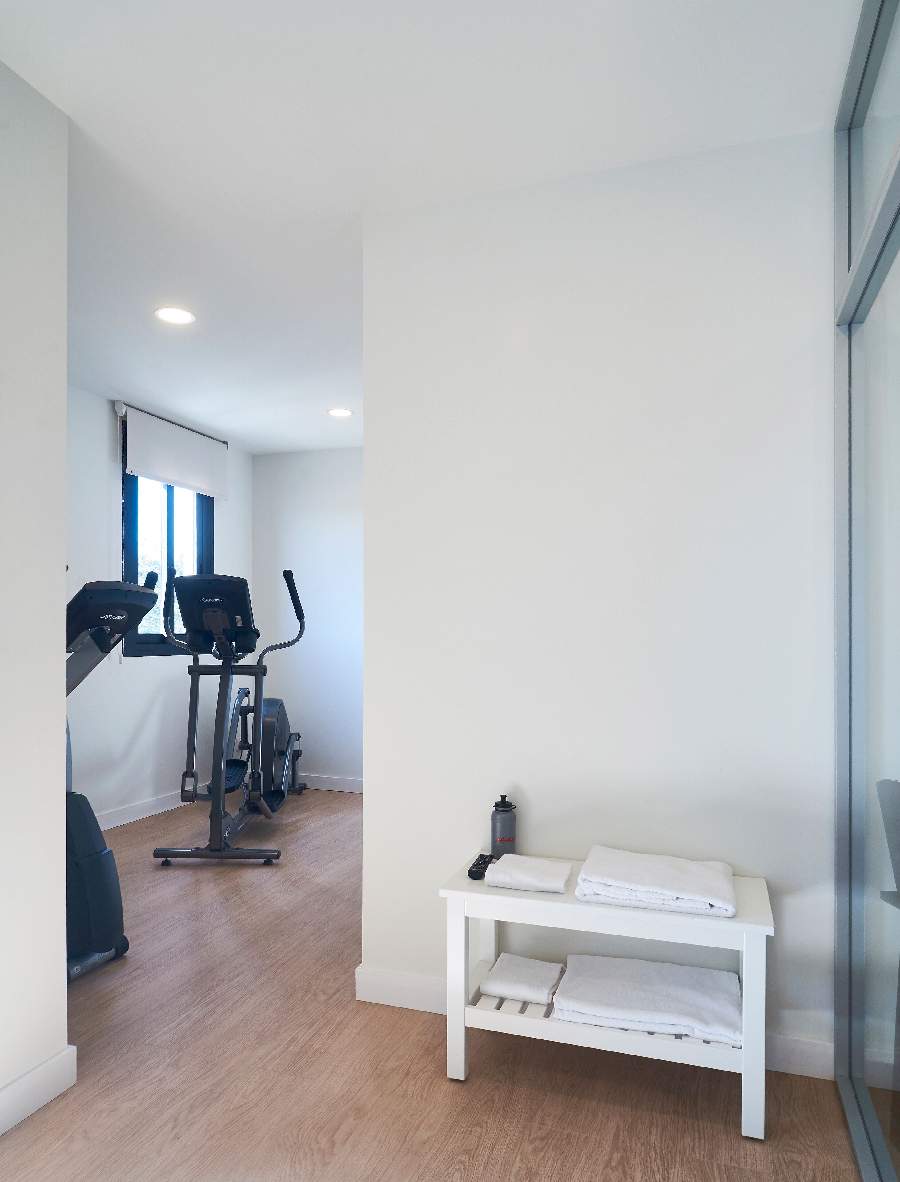 A modern, well-lit room with a treadmill, stationary bike, and a bench with towels.
