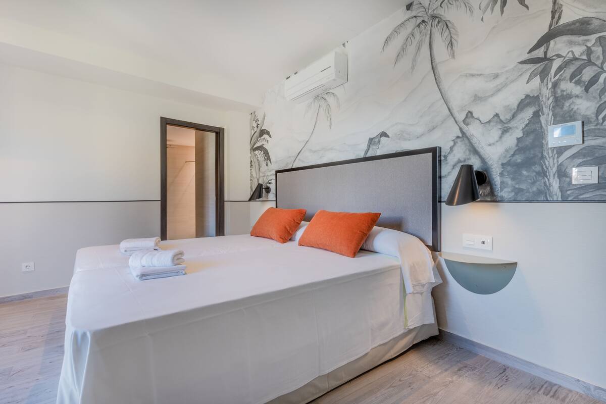 Une chambre d’hôtel moderne avec un grand lit, du linge de lit blanc, des oreillers orange, une tête de lit, une lampe fixée au mur et une fresque murale aux motifs tropicaux.