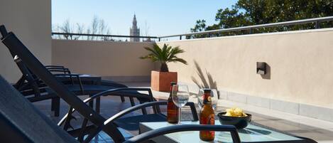 A rooftop terrace with lounge chairs, a table with a glass top, a potted plant, and bottles of beer.