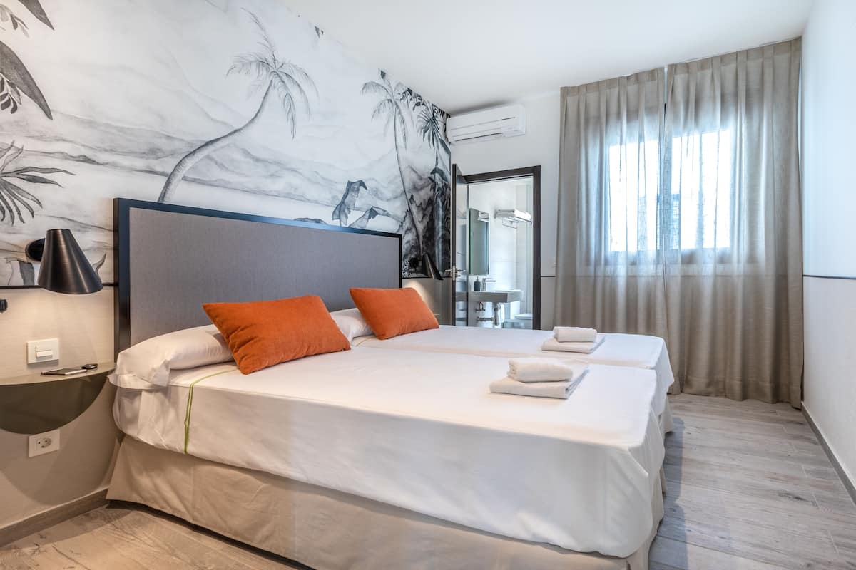 Une chambre d’hôtel avec deux lits, une grande fresque murale, un lavabo et une fenêtre avec des rideaux.