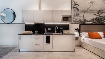 A compact kitchen with white cabinets, a microwave, and a sink. There is a bed with orange pillows to the right.