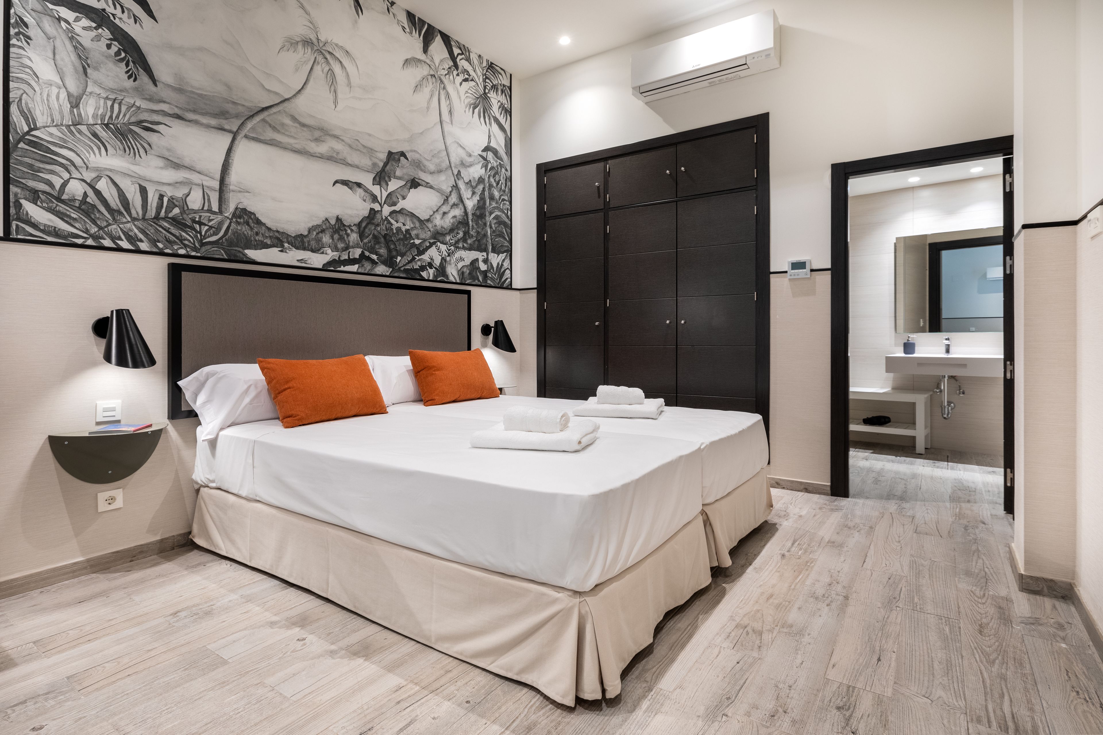 A double bed with white linens and orange pillows, a large dark wardrobe, a wall-mounted air conditioner, and a bathroom visible through an open door.