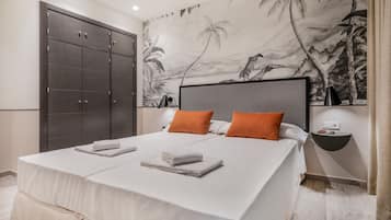 Una cama bien hecha con sábanas blancas y almohadas naranjas, un armario de madera oscura y un mural en la pared con paisaje tropical.