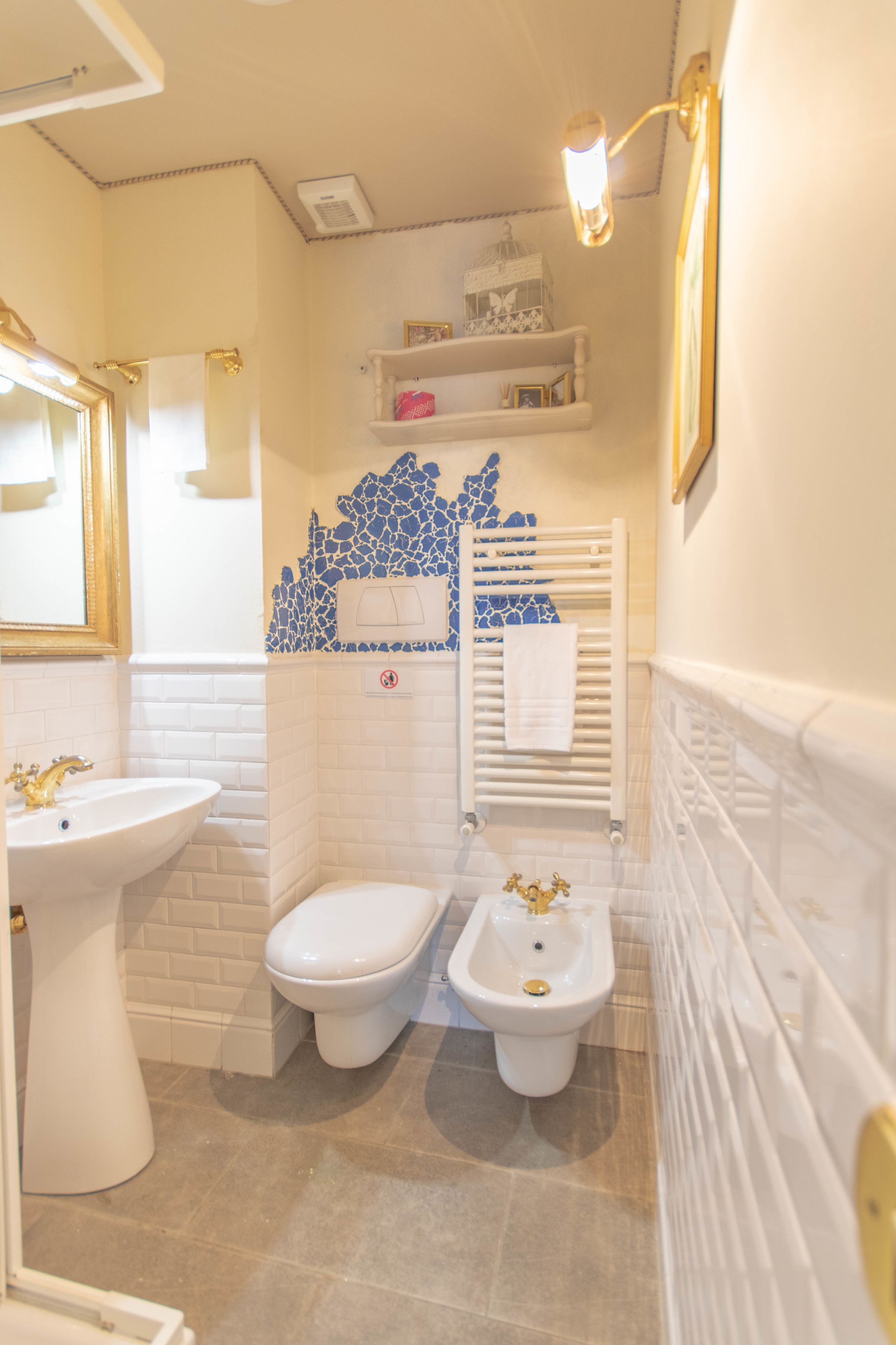 Une salle de bain avec un lavabo blanc, deux toilettes, un bidet, une douche aux carreaux de mosaïque bleus et un porte-serviettes blanc.