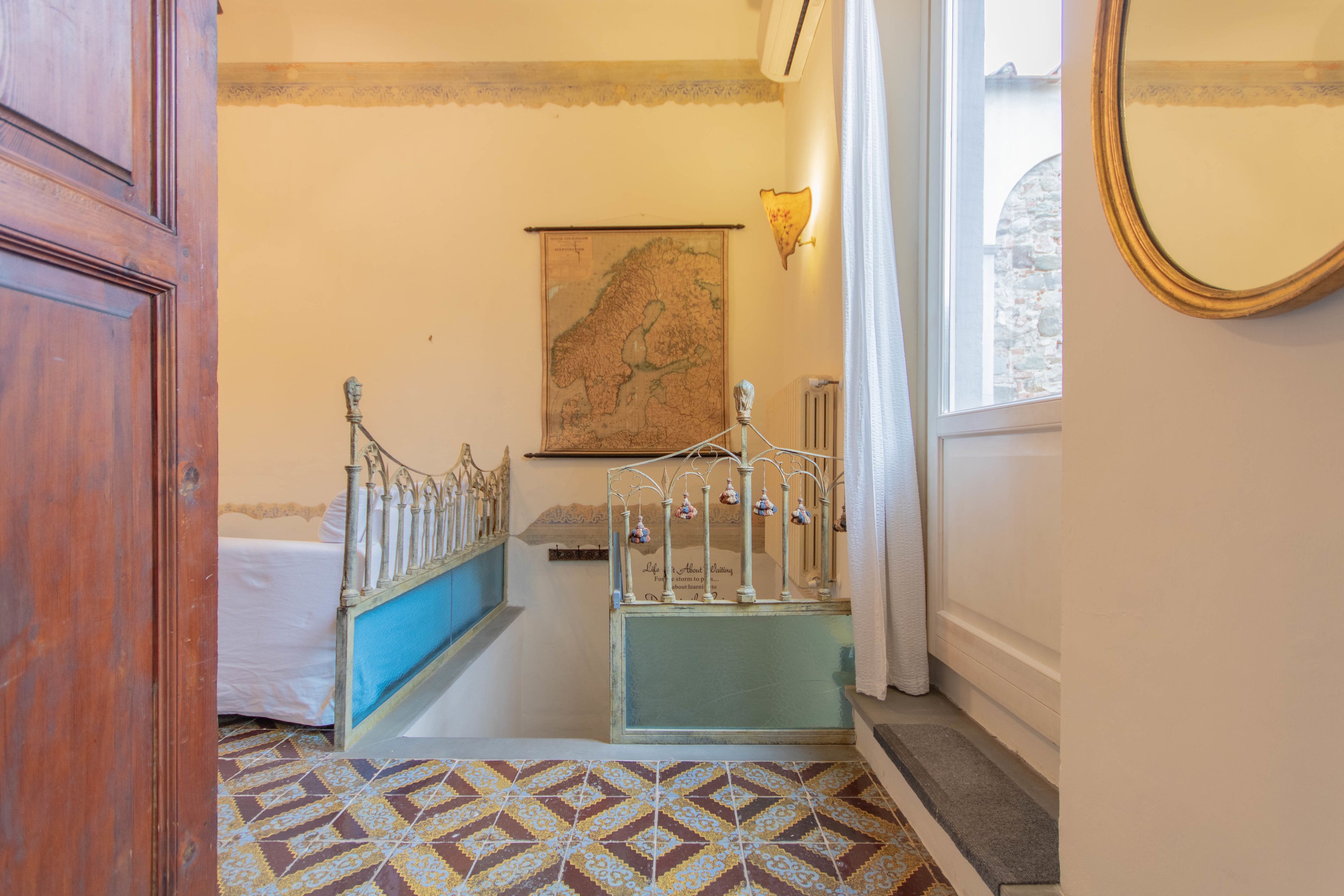 Un pasillo estrecho con suelo estampado, una cama con cabecera azul, un mapa en la pared y un espejo a la derecha.