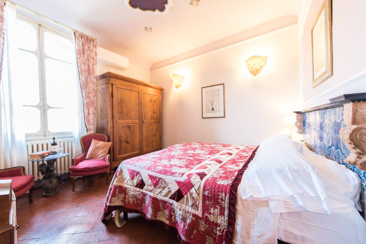 Een slaapkamer met een bed, een houten garderobe, een stoel, een tafel en een raam met gordijnen.
