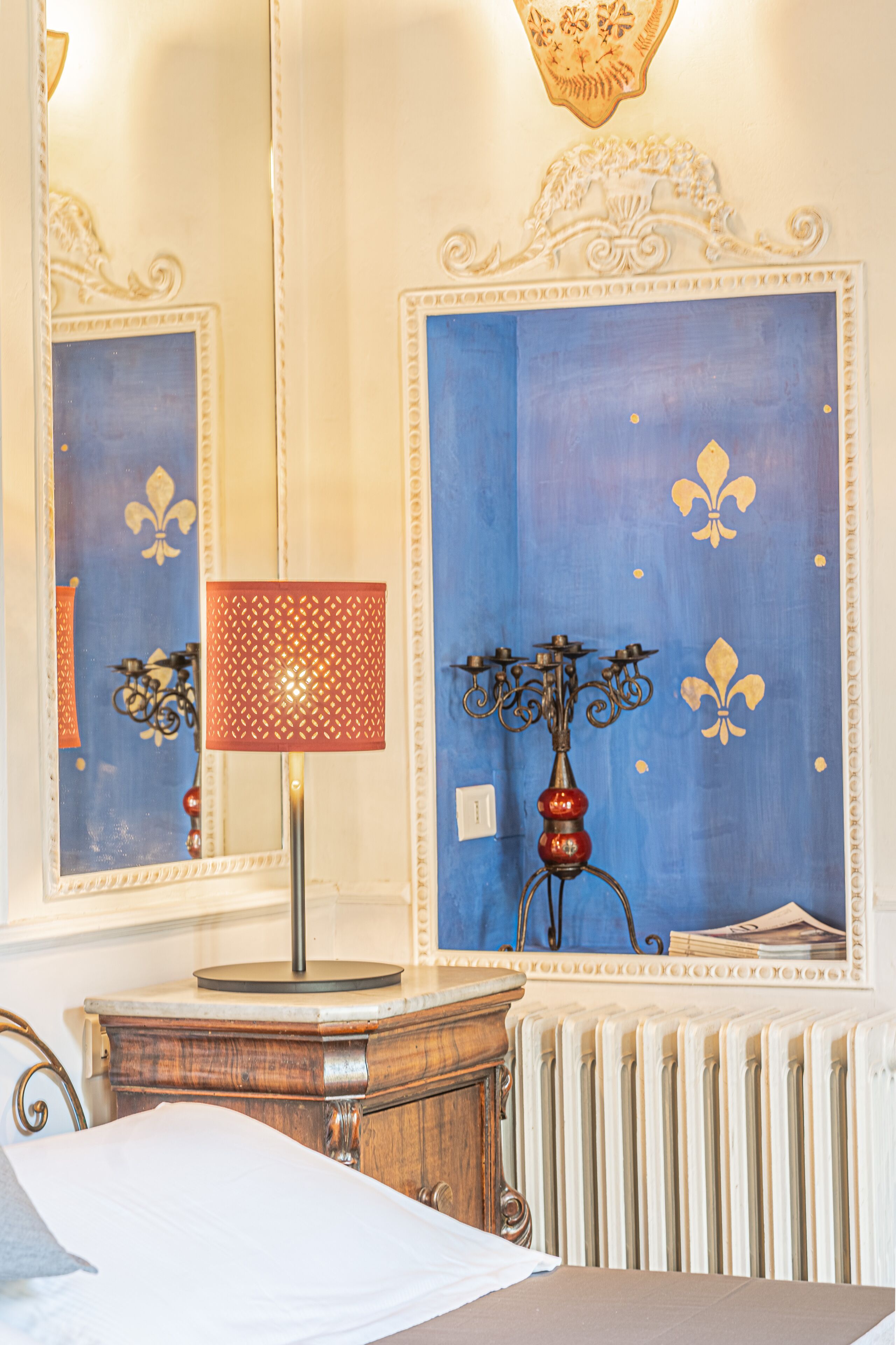 A room with a bed, a wooden dresser, a red patterned lamp, a candelabra, and a radiator.