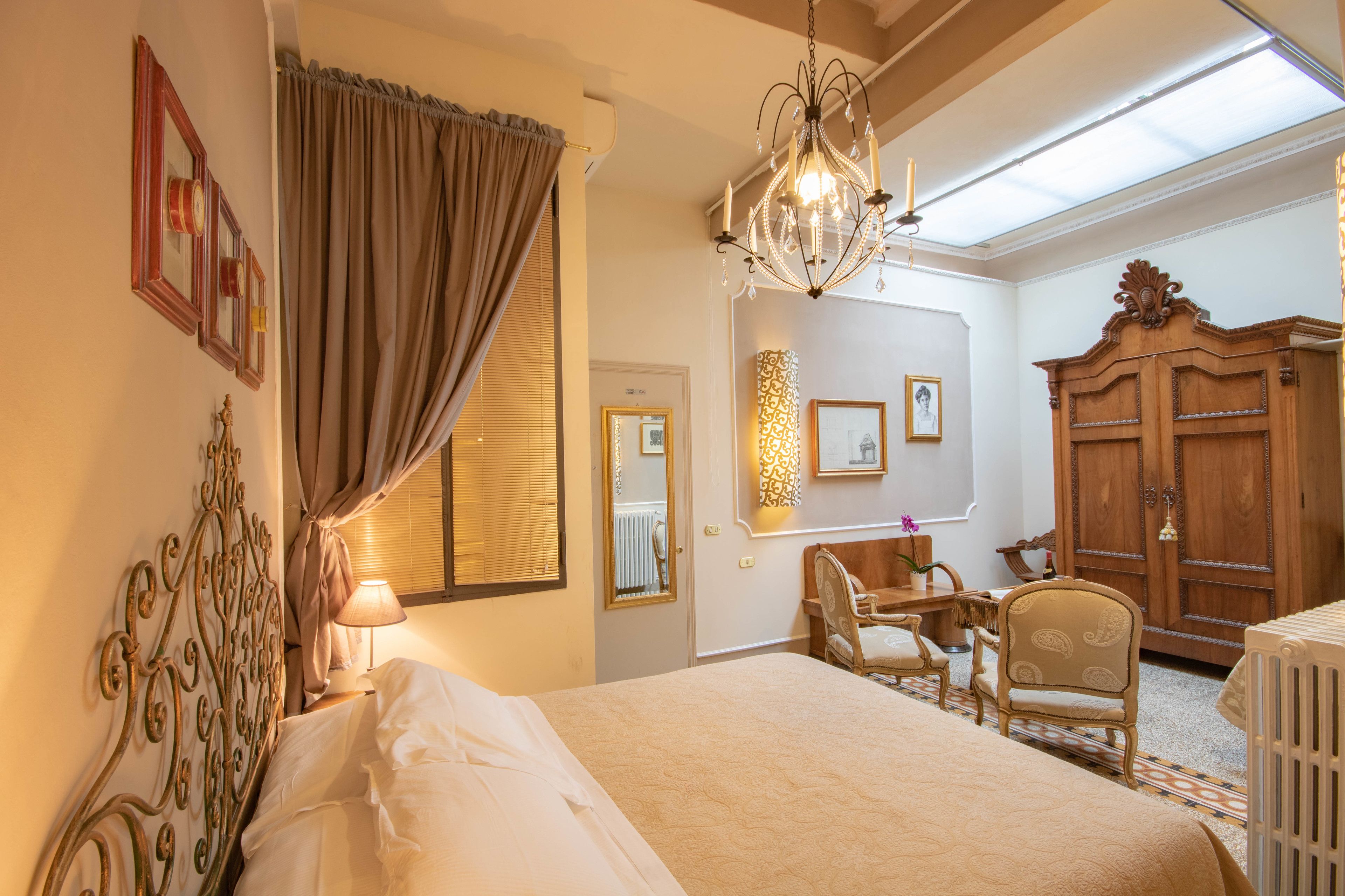 A hotel room with a large bed, a chandelier, a wooden wardrobe, and a desk with chairs.