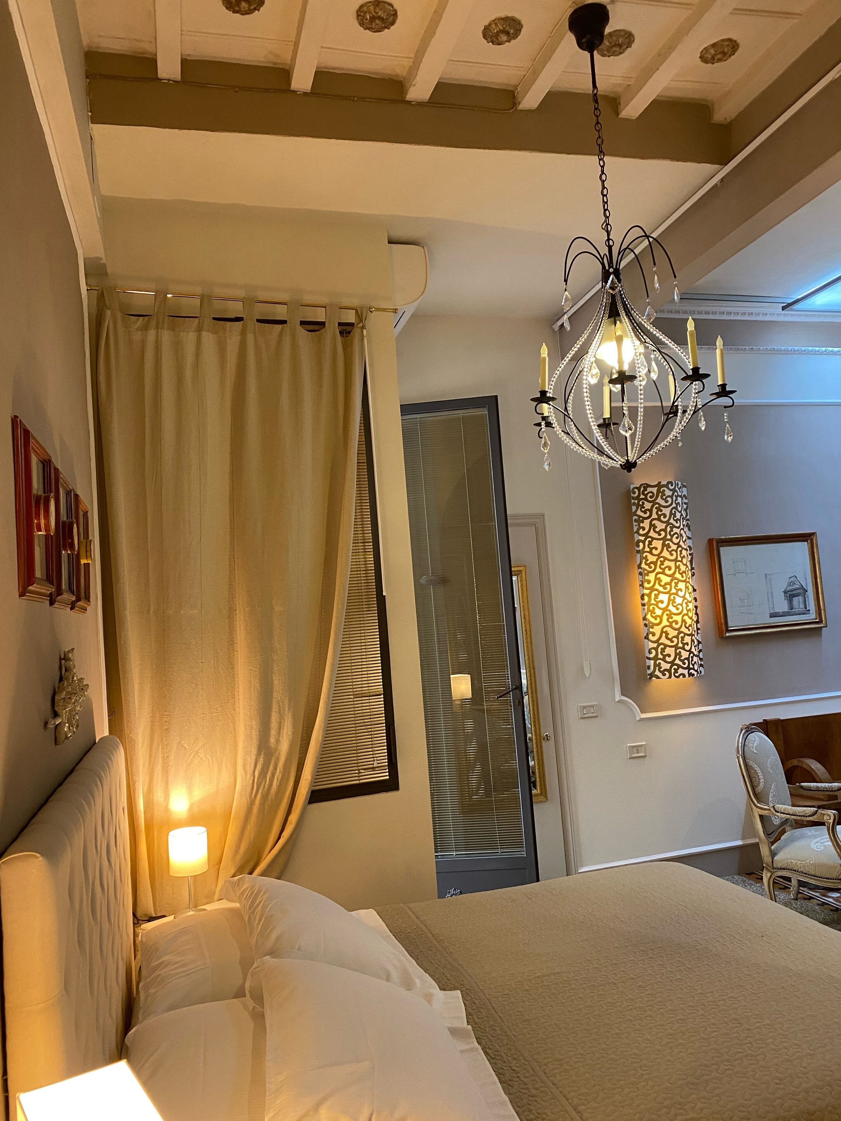 A bedroom with a large bed, a chandelier, a window with blinds, and a door with a mirror.