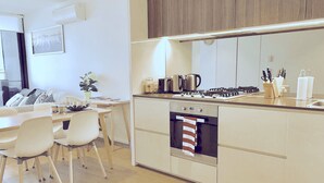 A modern kitchen with a built-in oven, a coffee maker, and a kettle. There is a dining area with a table and chairs. - Brilliant Victoria Harbour Waterfront (Docklands)