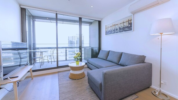 A modern living room with a grey sofa, a small round table with a vase, a floor lamp, and a large window offering a city view. - Brilliant Victoria Harbour Waterfront (Docklands)