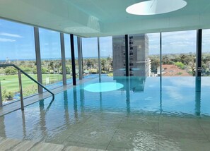 An indoor infinity pool with clear views of the surrounding landscape, including a golf course and distant buildings. - Fawkner Apartment 1B1B (Melbourne)