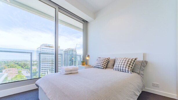 Una habitación de hotel moderna con una cama grande, almohadas a cuadros y vistas de la silueta urbana a través de ventanas de piso a techo.
