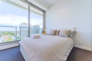 Quarto de hotel moderno com uma cama grande, almofadas xadrez e vista para o horizonte da cidade através de janelas do chão ao teto.