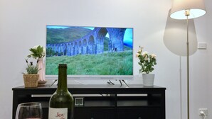 A living room with a flat-screen TV displaying a scenic landscape, a bottle of wine, a glass of wine, and two potted plants. - Fawkner Apartment Bay-view LXI (Melbourne)