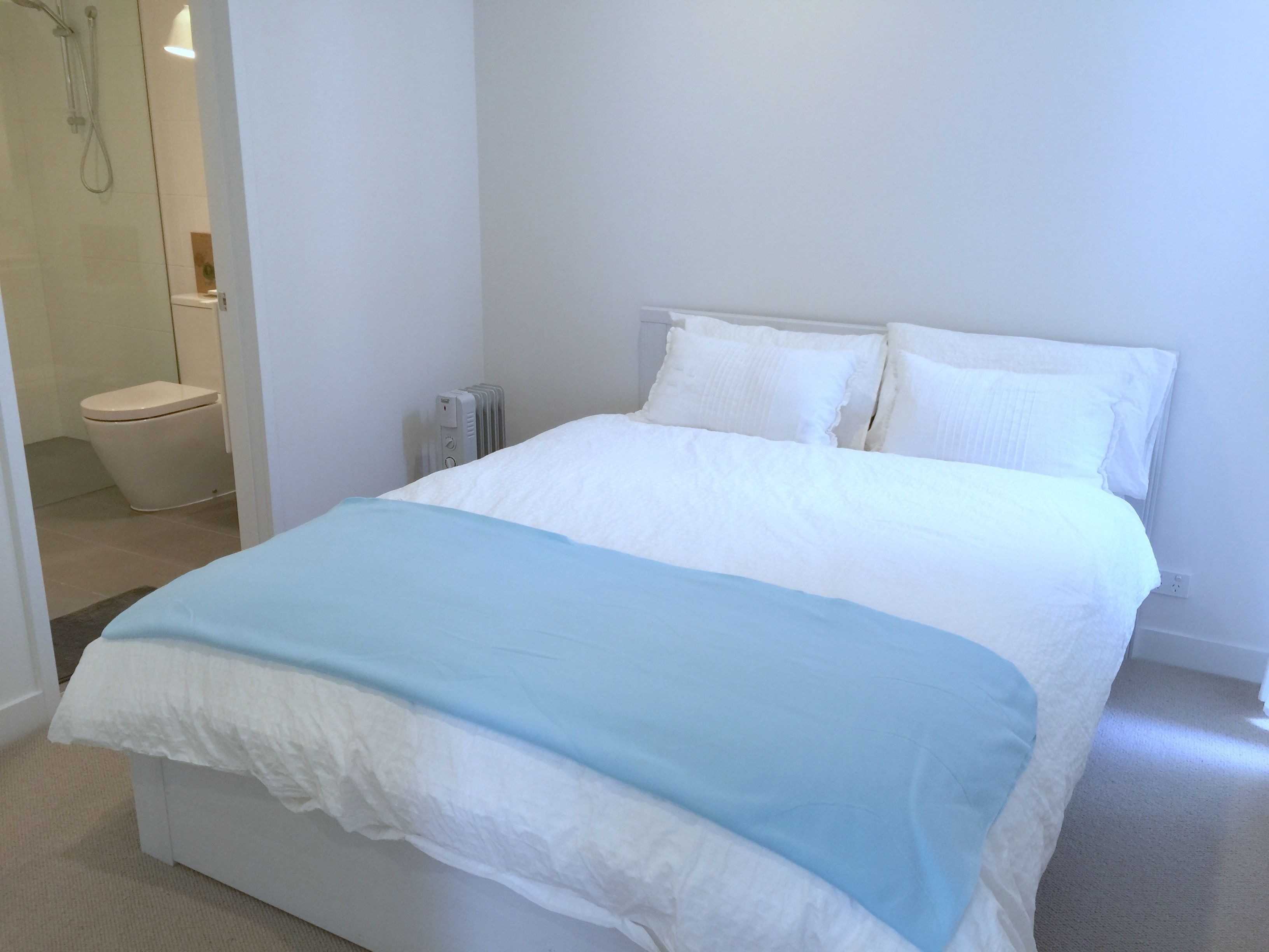 Un dormitorio con cama, ropa de cama blanca, un radiador y un inodoro visible en el baño contiguo.