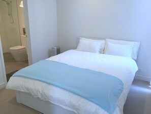 Quarto com cama, roupa de cama branca, radiador e casa de banho adjacente com sanita visível.