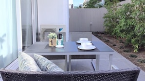 An outdoor patio with a table set for two, a wicker chair, and a small garden area with a fence and trees. - Brand New Stunning Home with Garden (Brighton)