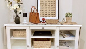 A white console table with a framed picture, a vase with white flowers, a brown handbag, a small red flower, a woven basket, and a potted plant. - Brand New Stunning Home with Garden (Brighton)