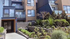 A modern multi-story residential building with a brick facade, large windows, and a landscaped garden area with various plants and a small water feature.