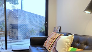 A modern living room with a black leather sofa, a glass coffee table, and a view of a brick building through large windows.