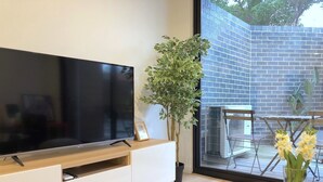A modern living room with a flat-screen TV on a wooden TV stand, a potted plant, and a glass table with flowers.