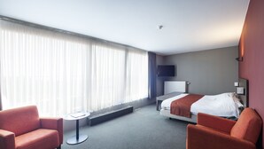 Une chambre d’hôtel avec un lit, deux fauteuils, une petite table, un téléviseur et une fenêtre avec des rideaux.