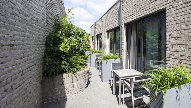 Un étroit couloir extérieur, avec un mur de briques d’un côté et un bâtiment moderne de l’autre. Près du bâtiment, on trouve une table et une chaise, ainsi qu’un petit espace jardin avec des plantes.