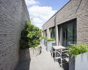 Un estrecho pasillo exterior con una pared de ladrillo en un lado y un edificio moderno en el otro. Cerca del edificio, hay un juego de mesa y silla, y un pequeño jardín con plantas.