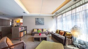 Une chambre d’hôtel moderne comprenant un coin salon, une table à manger, un ensemble de canapés, un coin cuisine et une grande fenêtre agrémentée de rideaux légers.